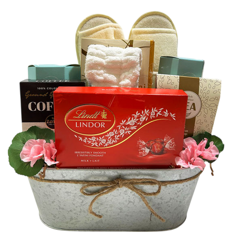Bliss gift basket featuring Lindt chocolate, gourmet coffee, tea, cozy slippers, and plush bath accessories, elegantly presented with floral accents and a rustic metal container.